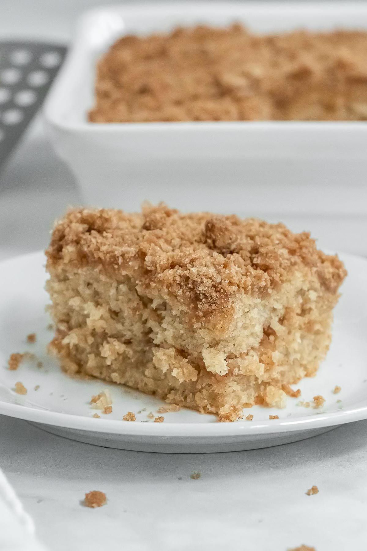 a slice of coffee cake with streusel topping on a white plate with the white baking dish in the background