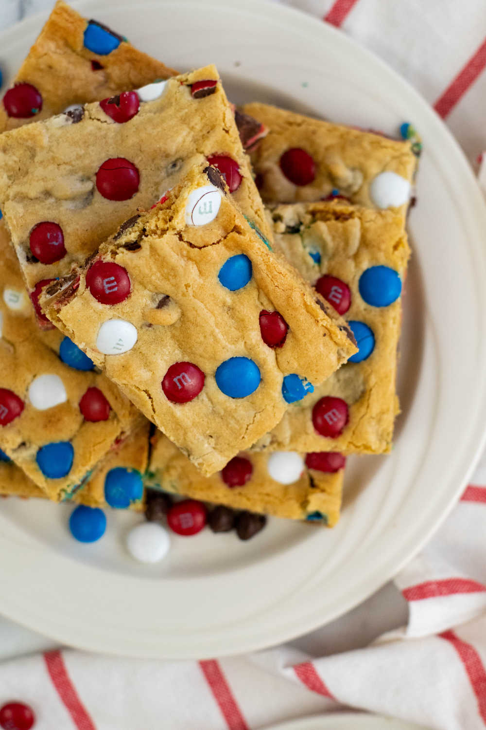A pile of red white and blue cookie bars on a white plate with a red striped linen. 