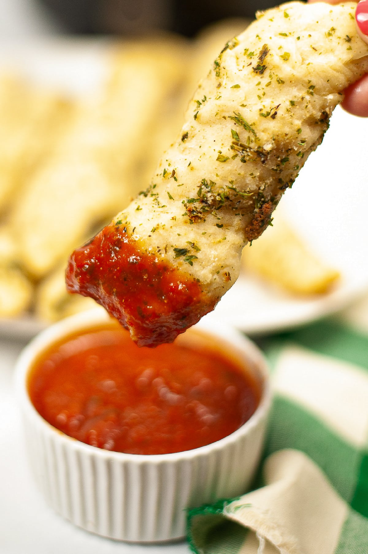 Close up side view of a breadstick covered in italian herbs being dipped in a small bowl of marinara with more breadsticks on a plate in the background.