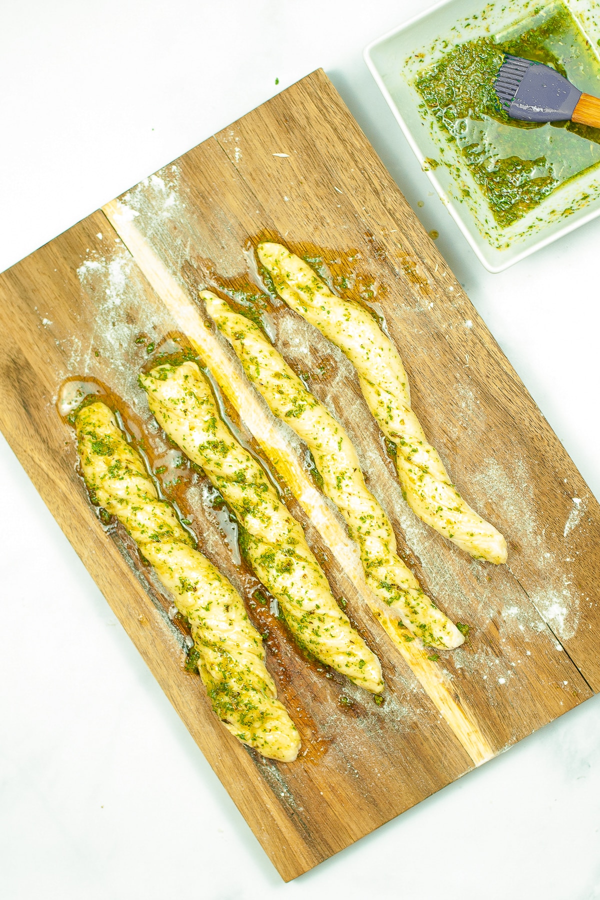 breadstick dough cut on a cutting board coated in butter and herbs with a small bowl nearby on the counter with more melted butter, italian herbs and a silicone brush.