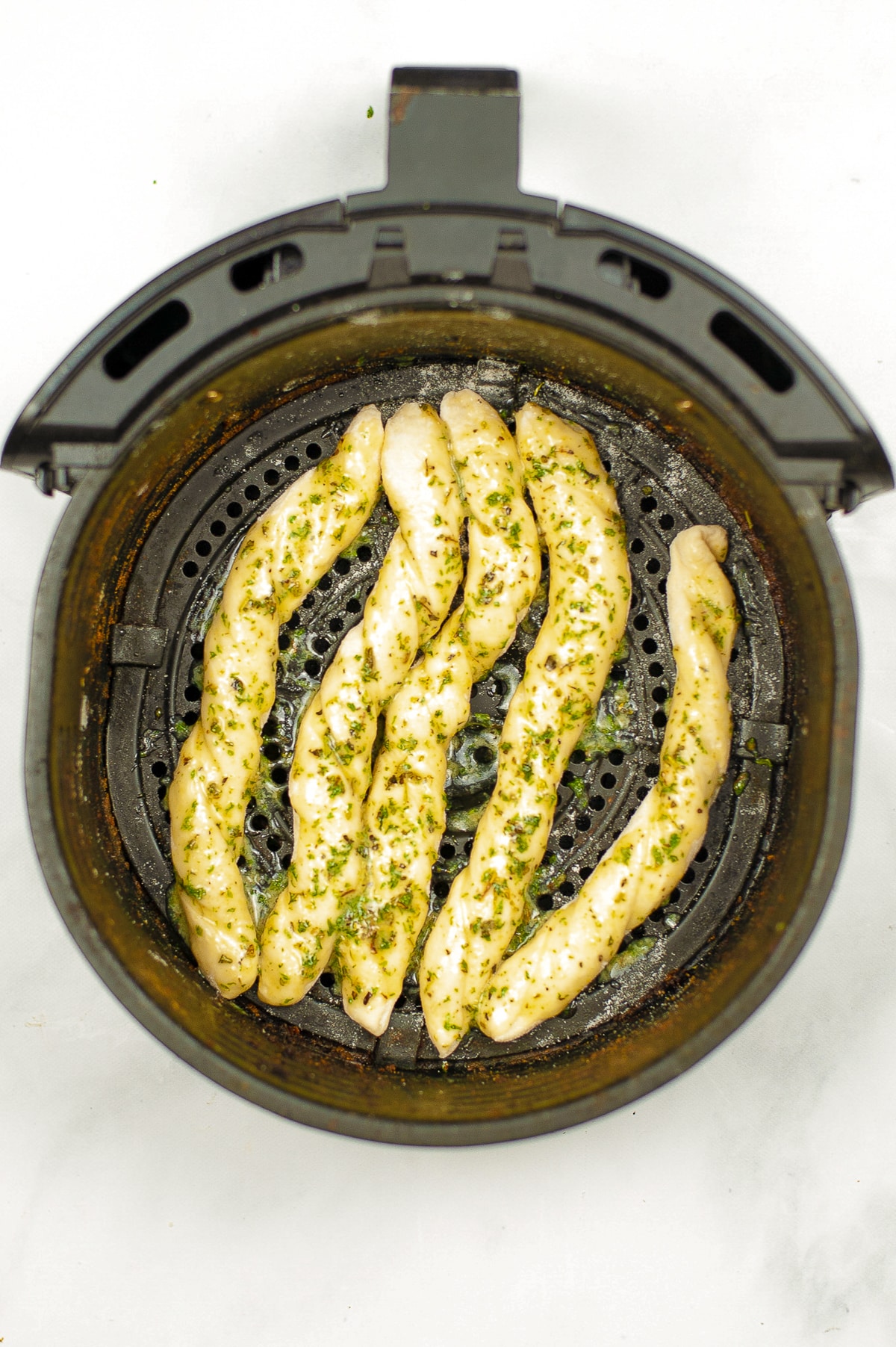 Raw breadstick dough cut into strips covered in butter and herbs in an air fryer basket from overhead.