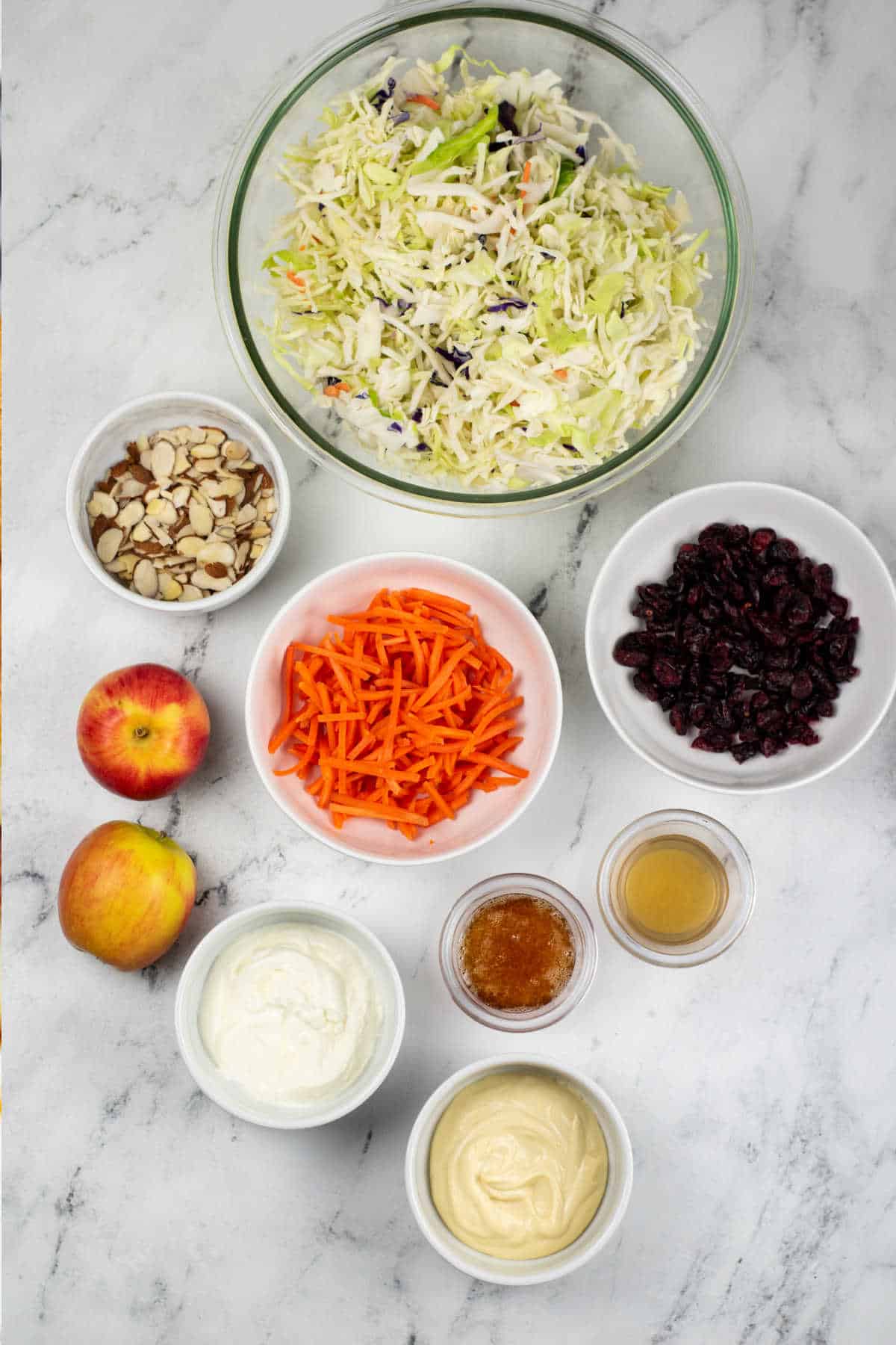 The ingredients for apple cranberry coleslaw on a white marble background. Coleslaw mix, sliced almonds, dried cranberries, shredded carrots, apples, honey, apple cider vinegar, sour cream and mayonnaise. 