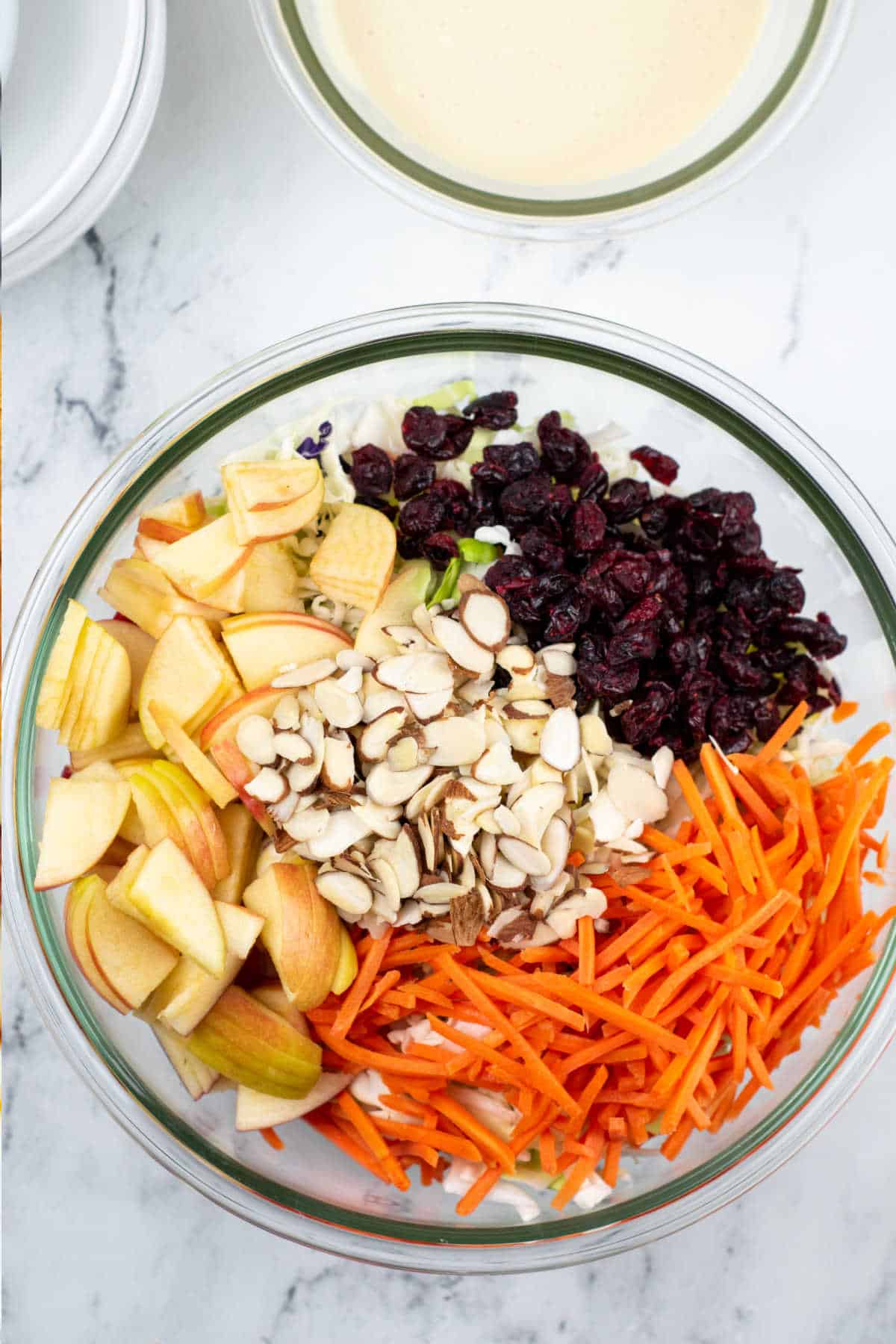 A large glass bowl with coleslaw mix, chopped apples, shredded carrots, sliced almonds, and dried cranberries in it. 