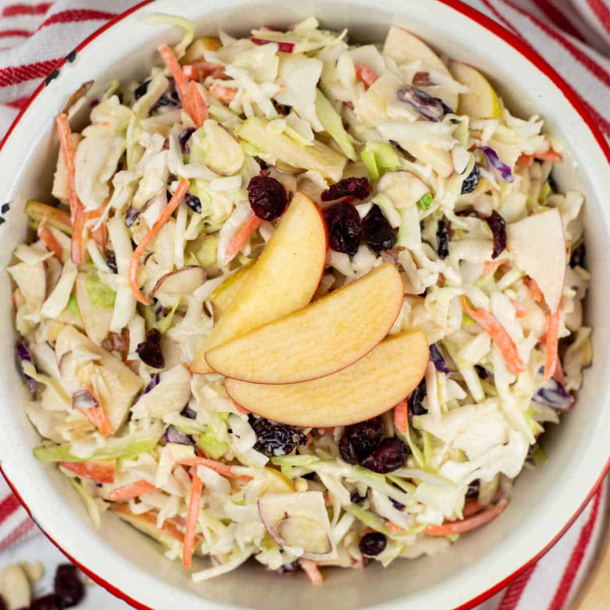 A white bowl full of coleslaw garnished with a sliced apple and dried cranberries. 