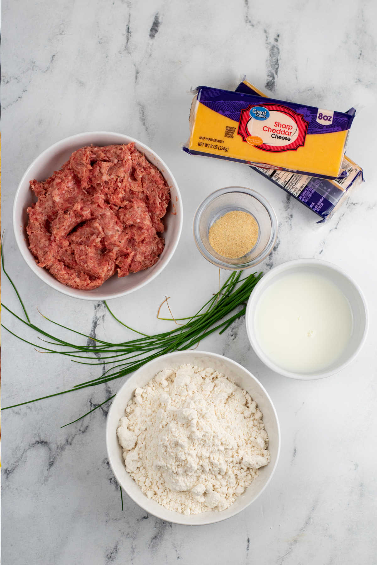 The ingredients for sausage cheese balls on a white marble background. Sausage, sharp cheddar cheese, garlic powder, fresh chives, milk and bisquick. 
