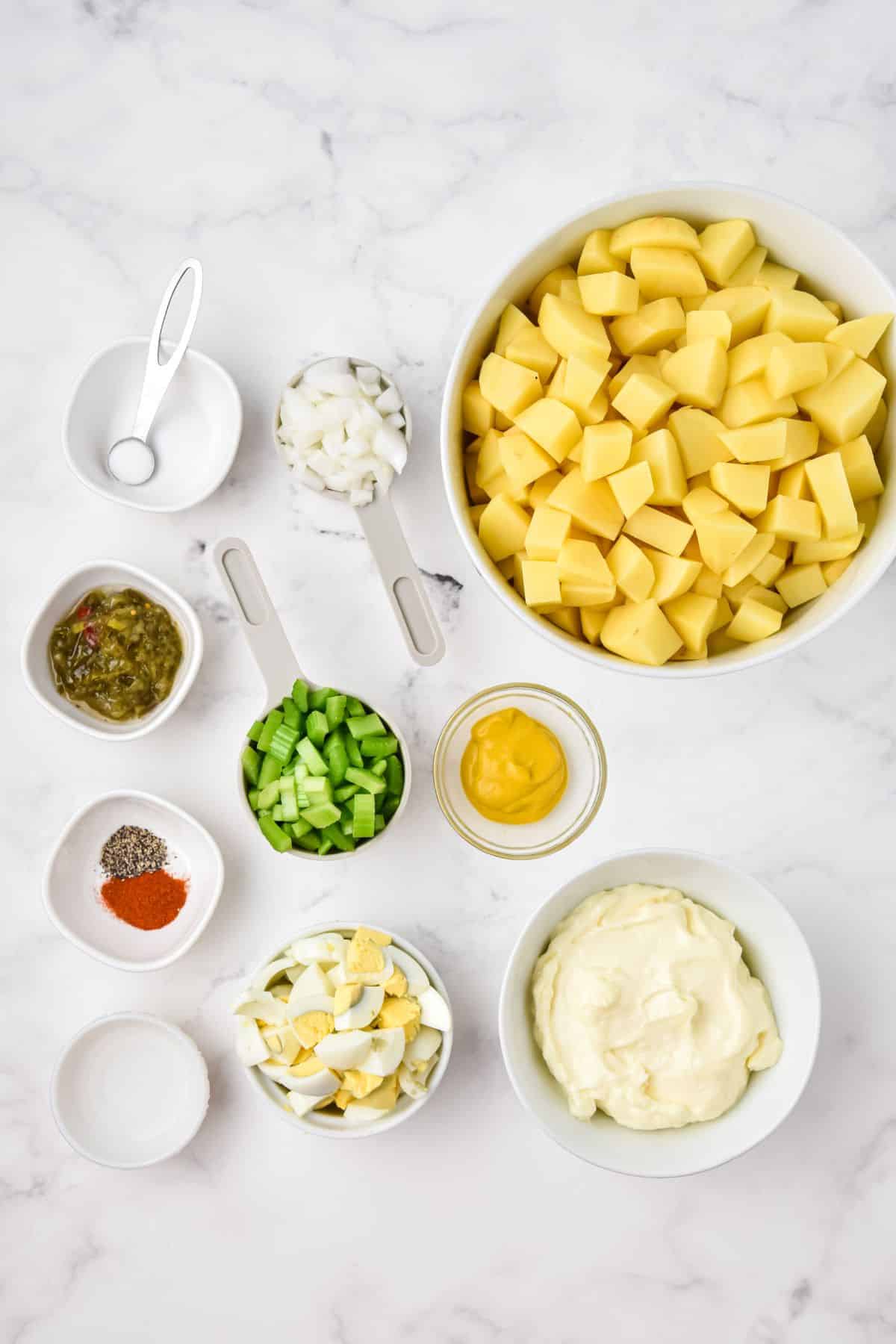ingredients in bowls on white counter: diced and peeled potatoes, mustard, chopped onions, salt, sweet pickle relish, chopped celery, pepper and paprika, vinegar, chopped hard boiled eggs, and mayonnaise