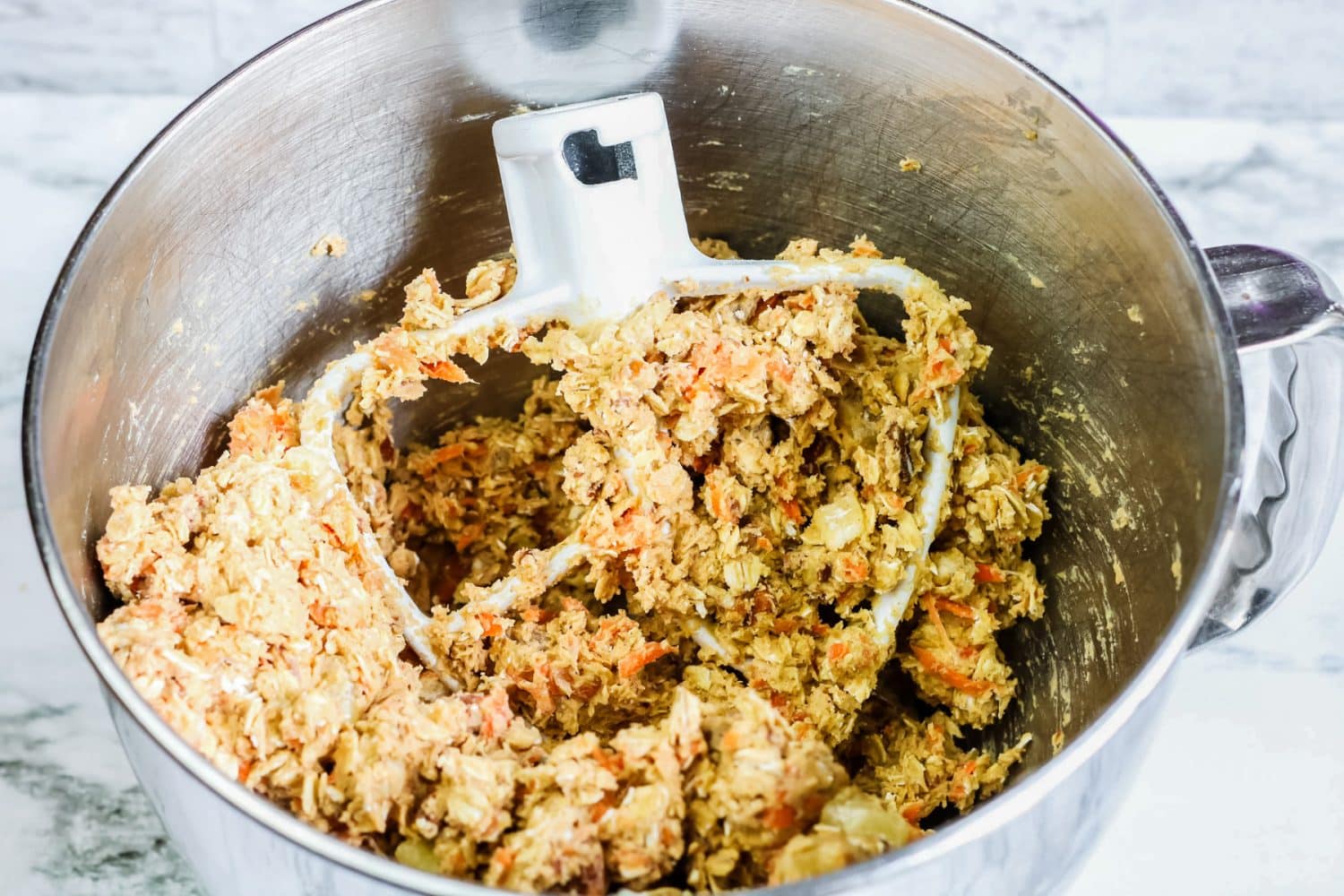 Batter for carrot oat cookies in a mixing bowl with an electric mixer paddle in the dough.