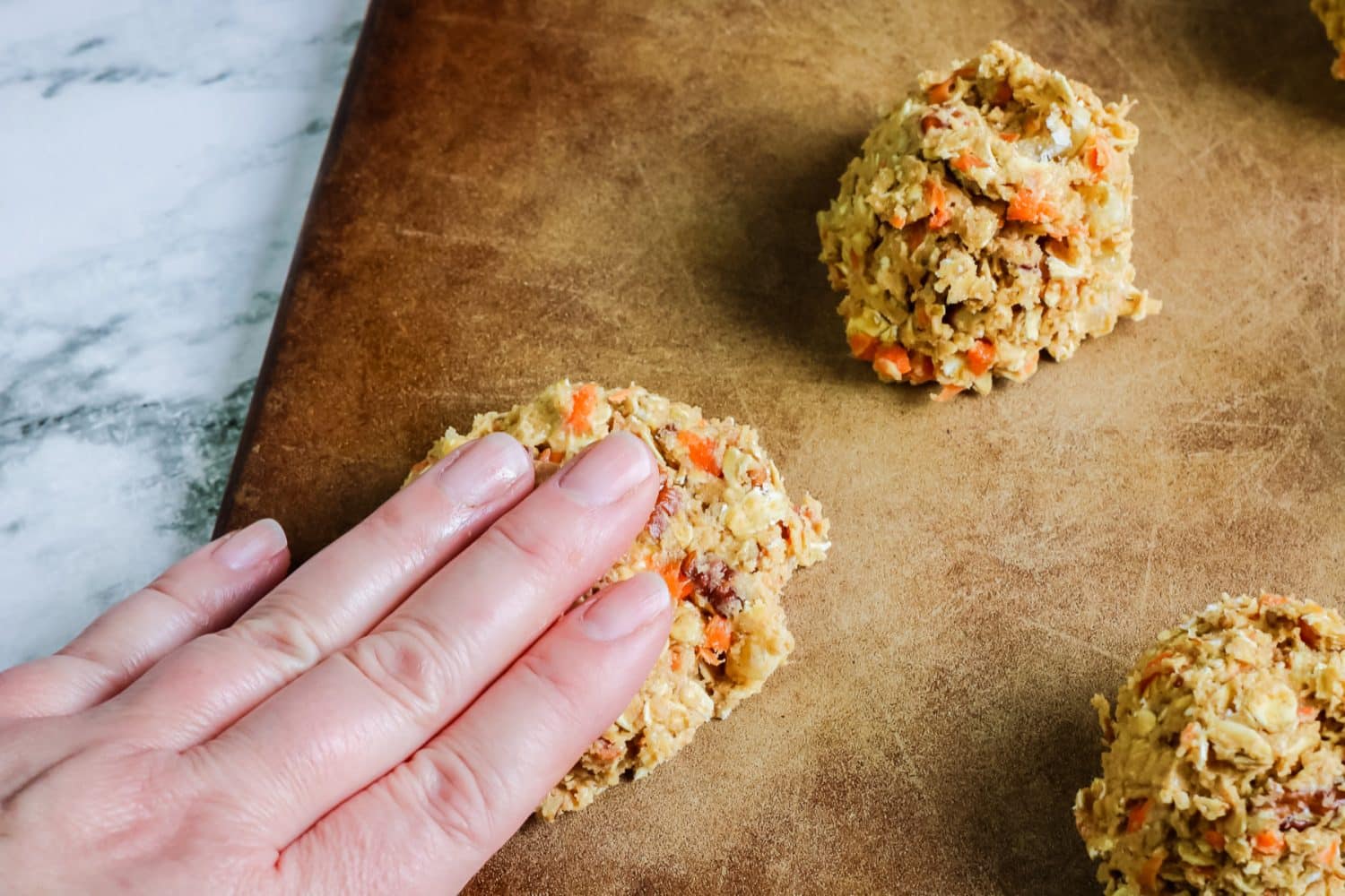 A person's fingers pressing carrot cookie dough balls on a baking sheet.