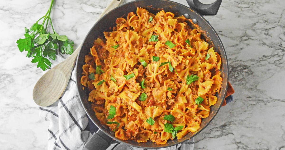 black skillet with a skillet lasagna recipe with bowtie pasta and ground beef in a tomato based sauce and garnished with fresh parsley.