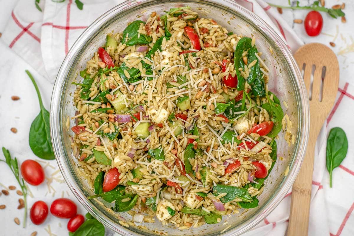orzo pesto salad recipe in a large glass bowl, surrounded by fresh spinach and tomatoes on a red-checked towel.