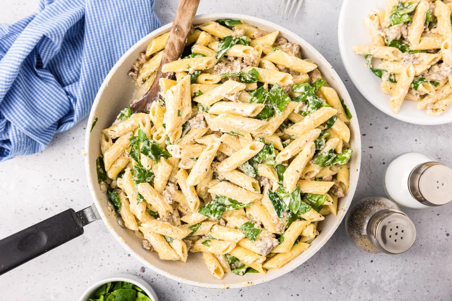 Creamy sausage pasta with spainch in a skillet being stirred by a wooden spoon.