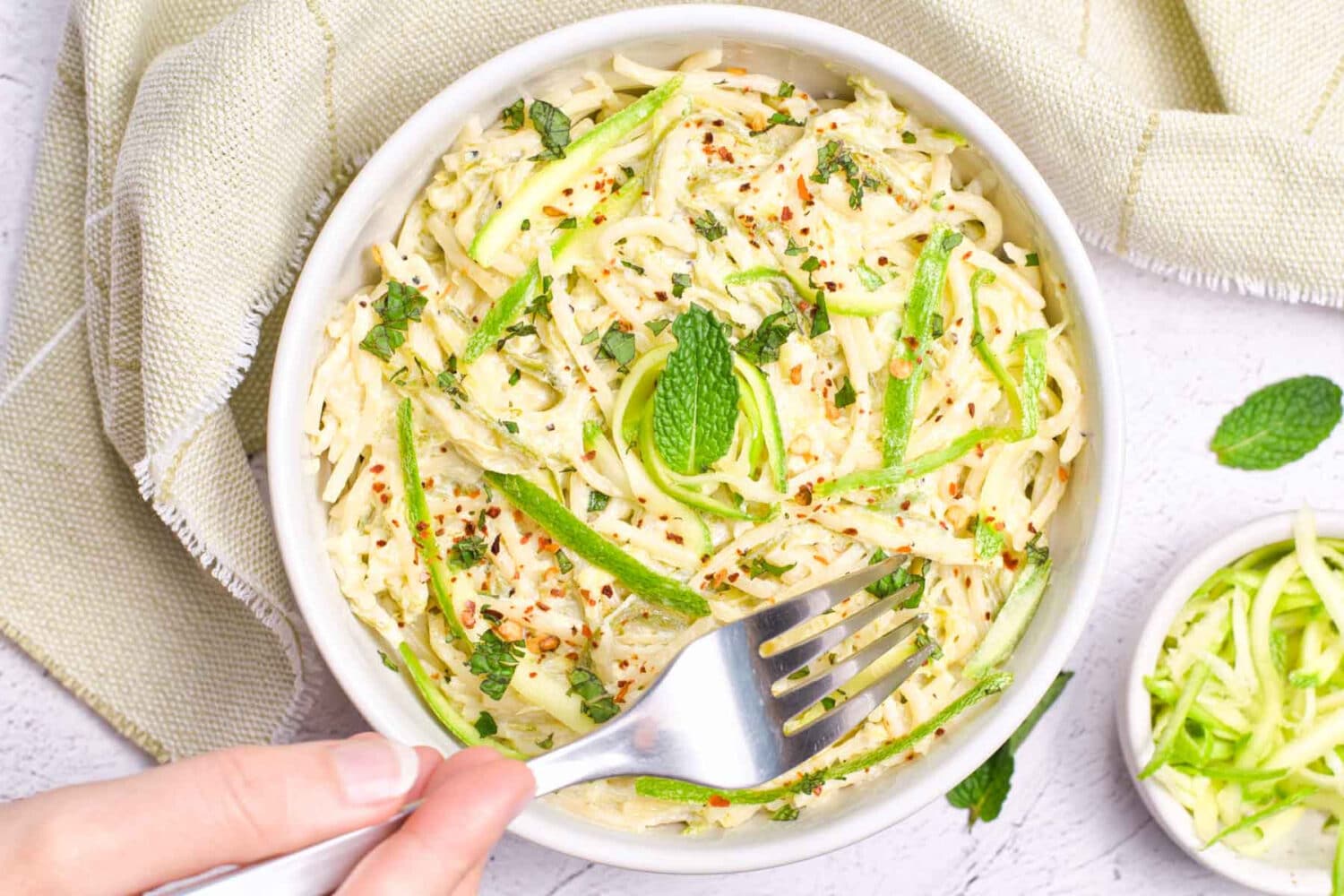 Pasta mixed with shredded zucchini, red peppet flakes and basil while a hand grabs a bite with a fork.
